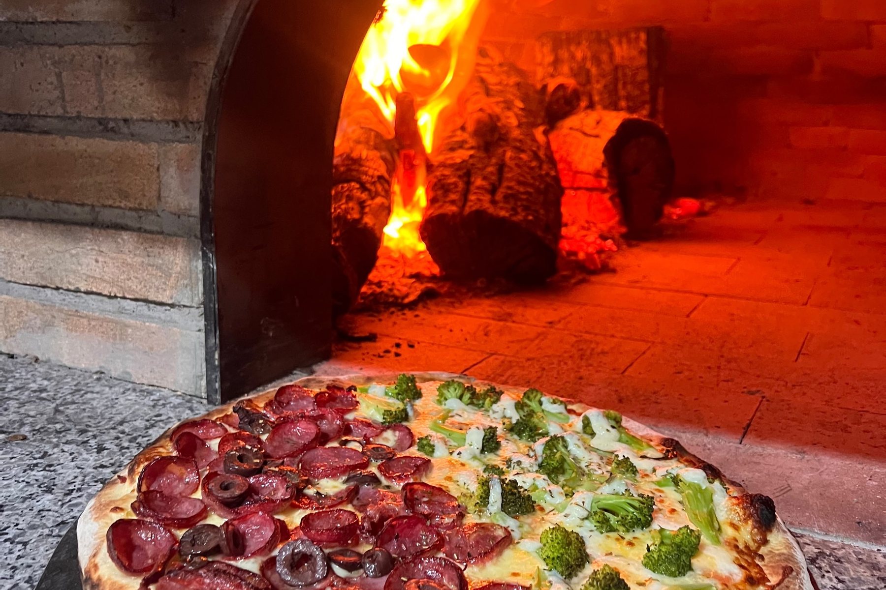 Chamas altas dentro do forno a lenha, com uma pizza meia calabresa meia brócolis na boca do forno